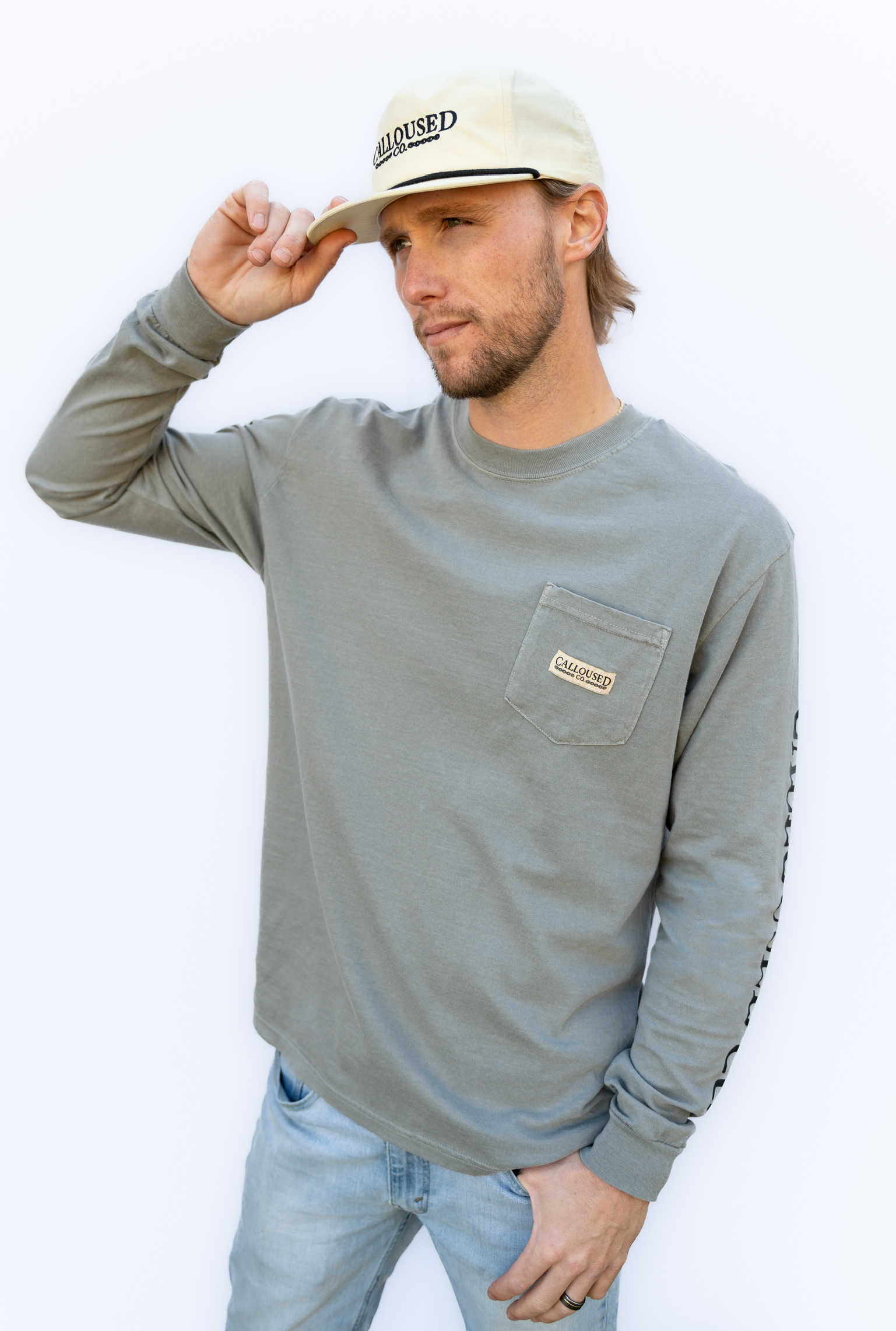Man wearing a gray 'Calloused Co' long-sleeve shirt with a pocket and a beige cap on a white background