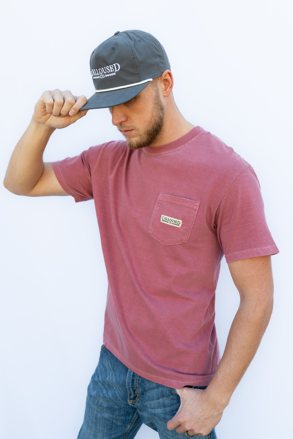 Man wearing a maroon 'Calloused Co' t-shirt with a pocket and a blue cap on a white background