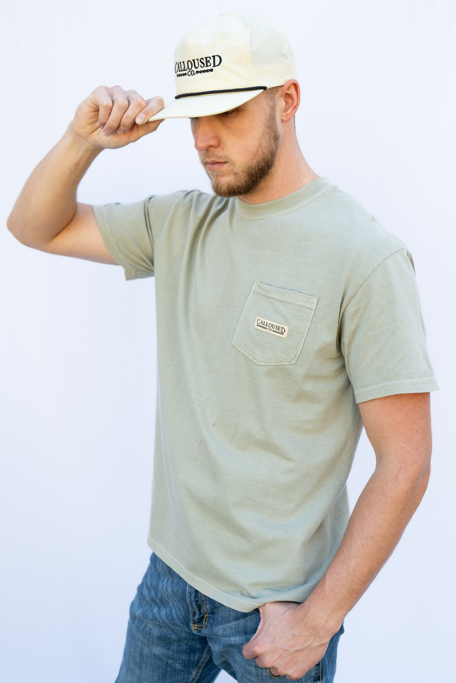 Man wearing a sandstone 'Calloused Co' t-shirt and beige cap with, posing against a white background