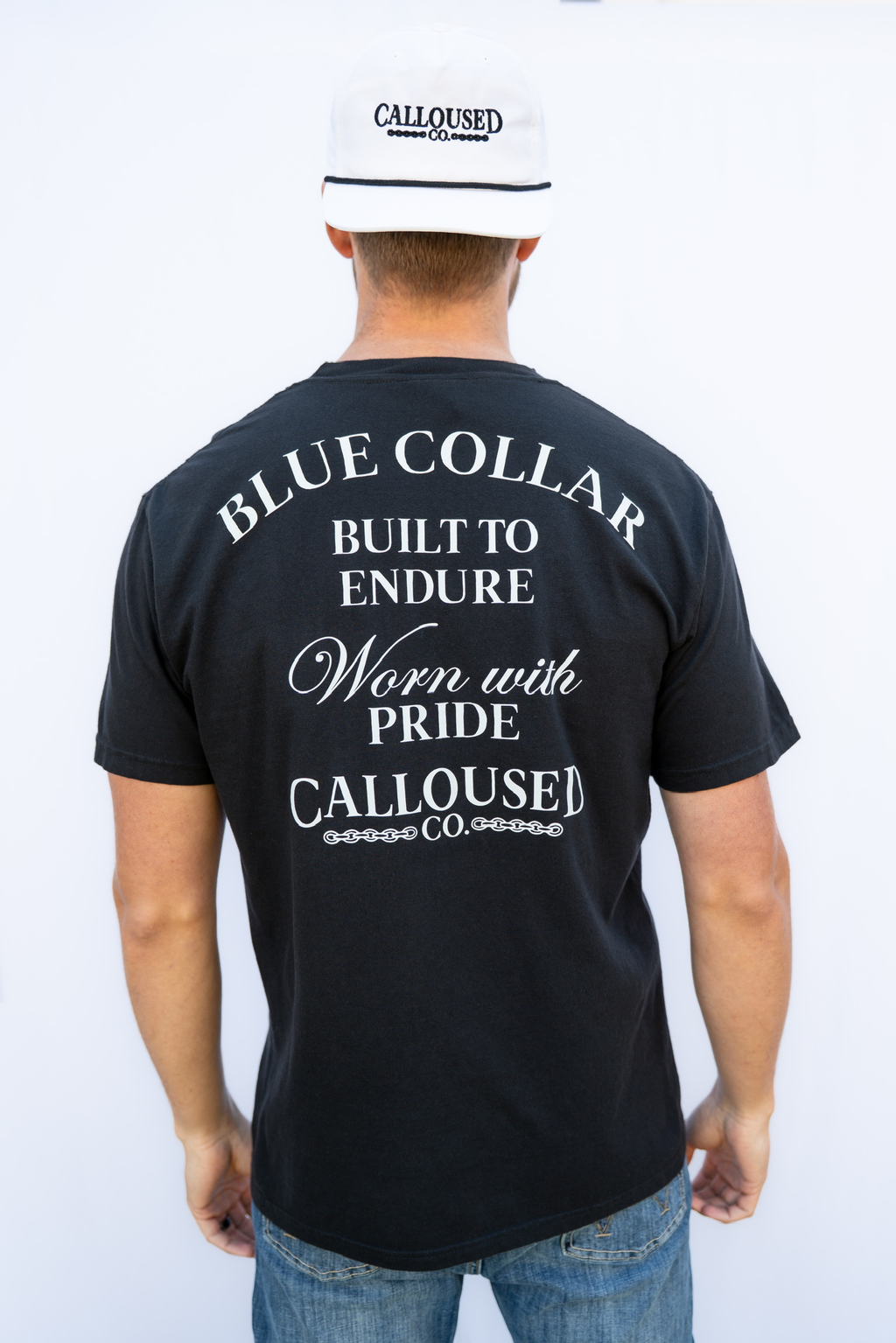 Person wearing a black t-shirt with white'Blue Collar Built to Endure Worn with Pride' text, 'Calloused Co' logo, and a white cap with 'Calloused Co' branding.