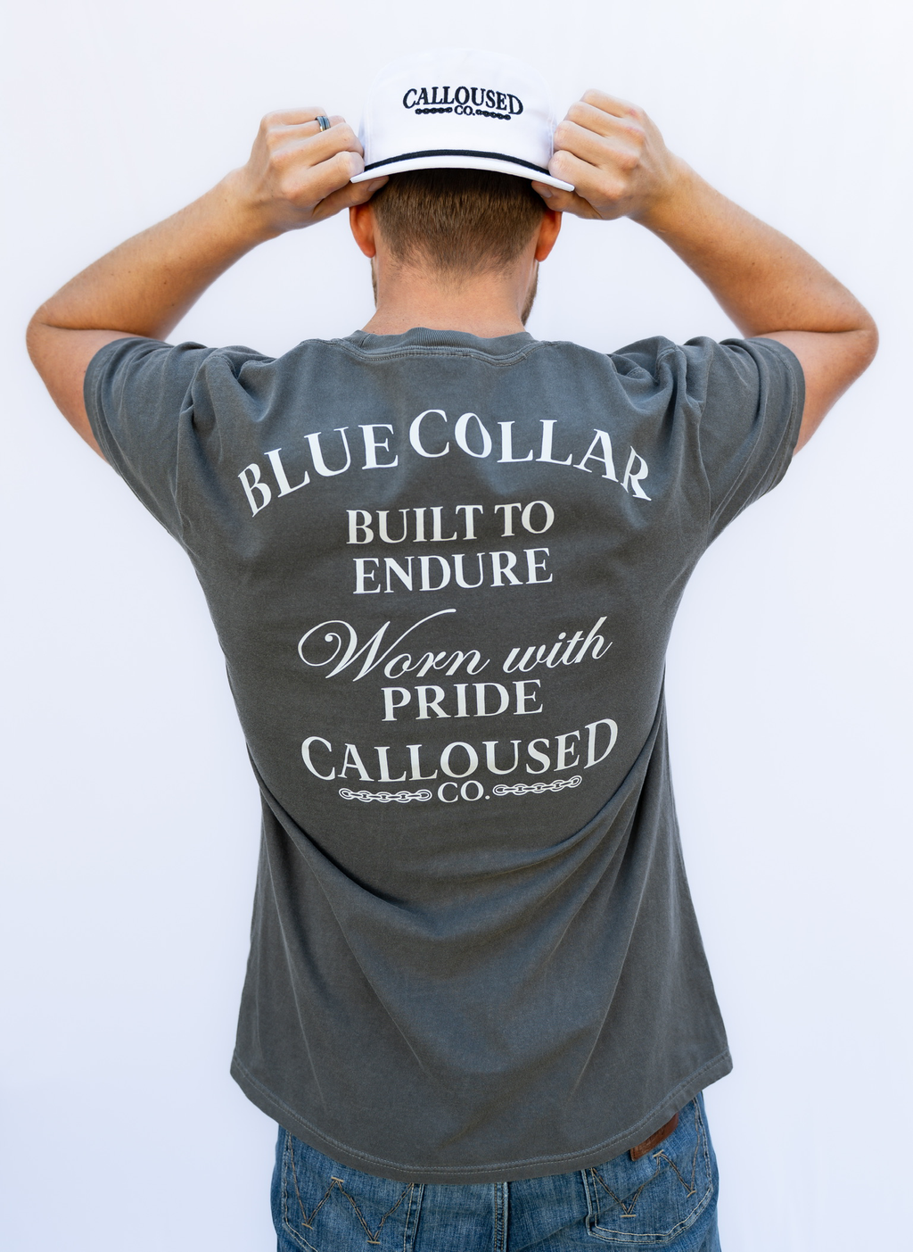 Person wearing a gray t-shirt with 'Blue Collar Built to Endure Worn with Pride' text, 'Calloused Co' logo, and a white cap, on a plain background