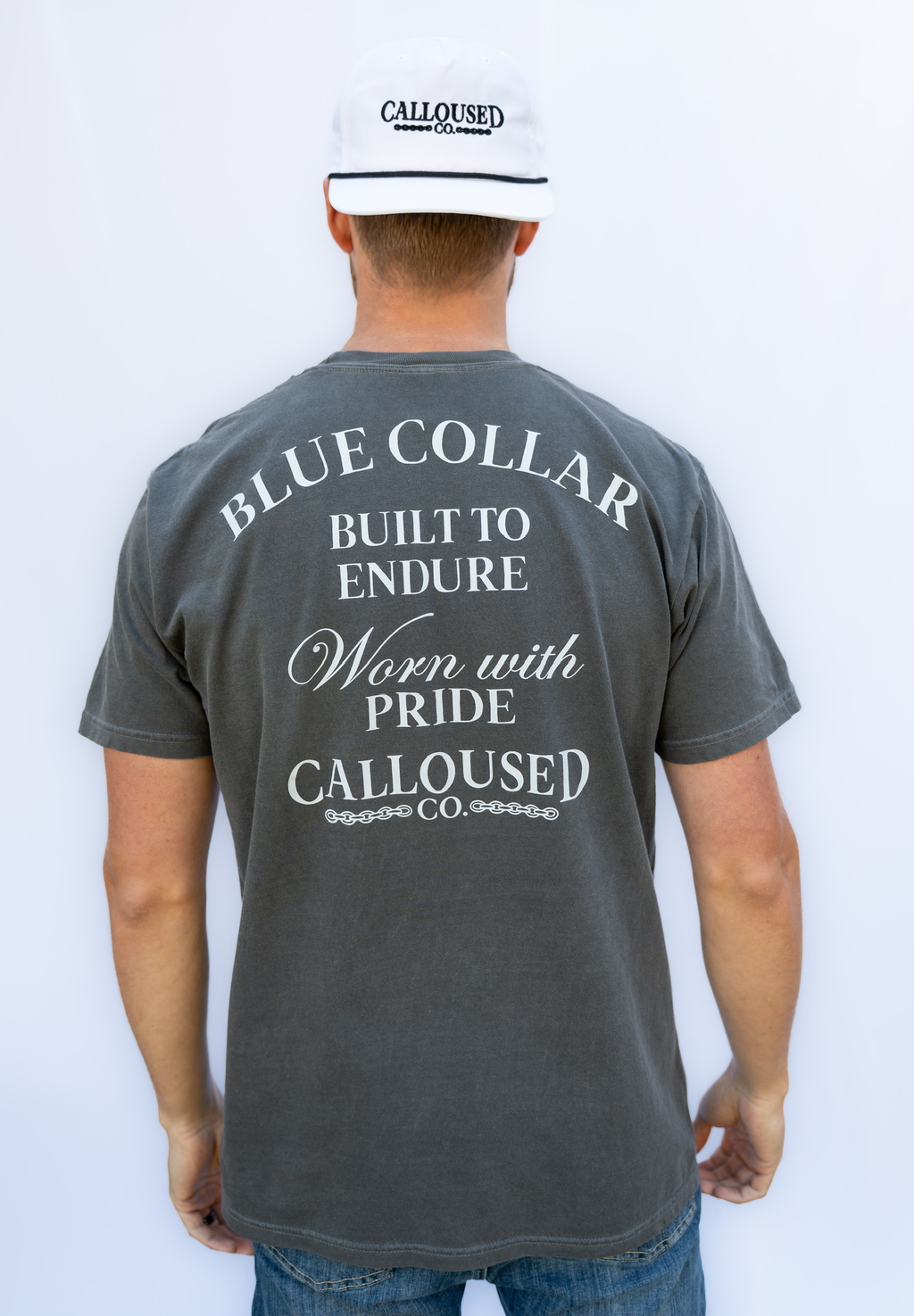 Person wearing a gray t-shirt with 'Blue Collar Built to Endure Worn with Pride' text, 'Calloused Co' logo on the back, standing against a white background