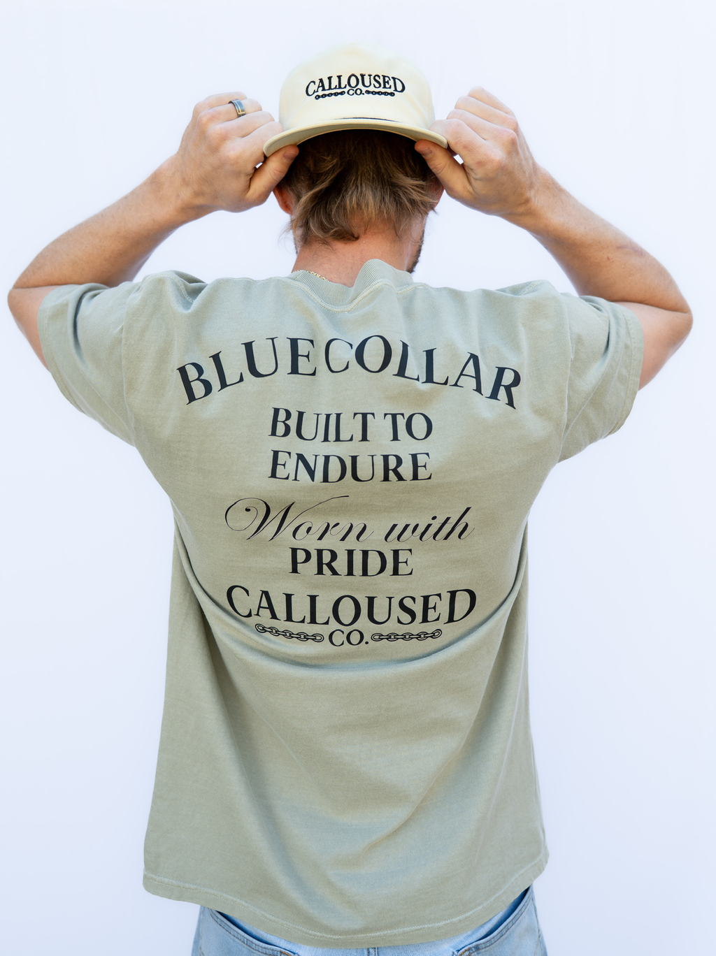 Person wearing a sandstone t-shirt with 'Blue Collar Built to Endure Worn with Pride' text, 'Calloused Co' logo, and a cap, on a plain background