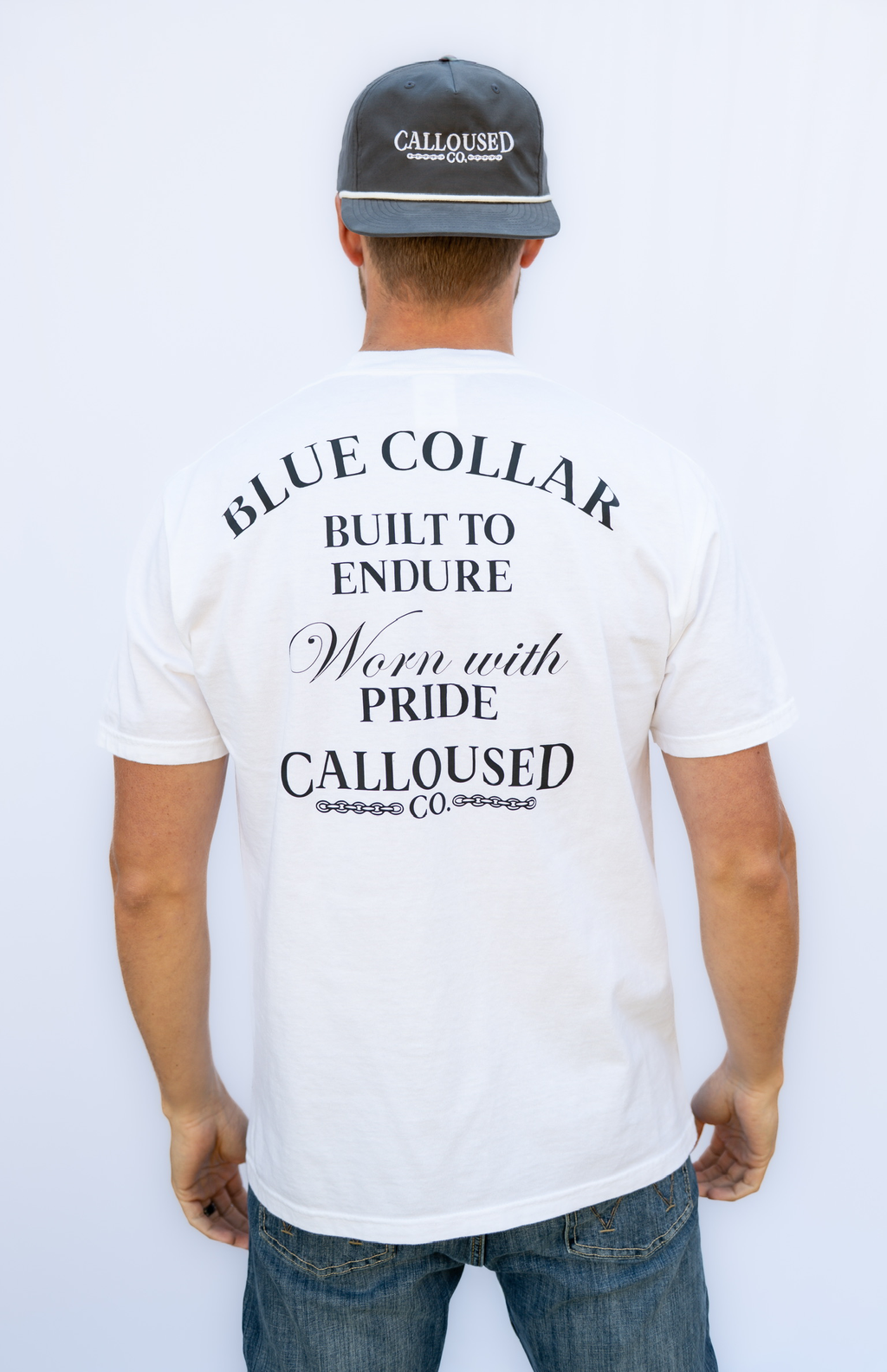Man wearing a white t-shirt with 'Blue Collar Built to Endure Worn with Pride' text, 'Calloused Co' logo, and a cap on a white background