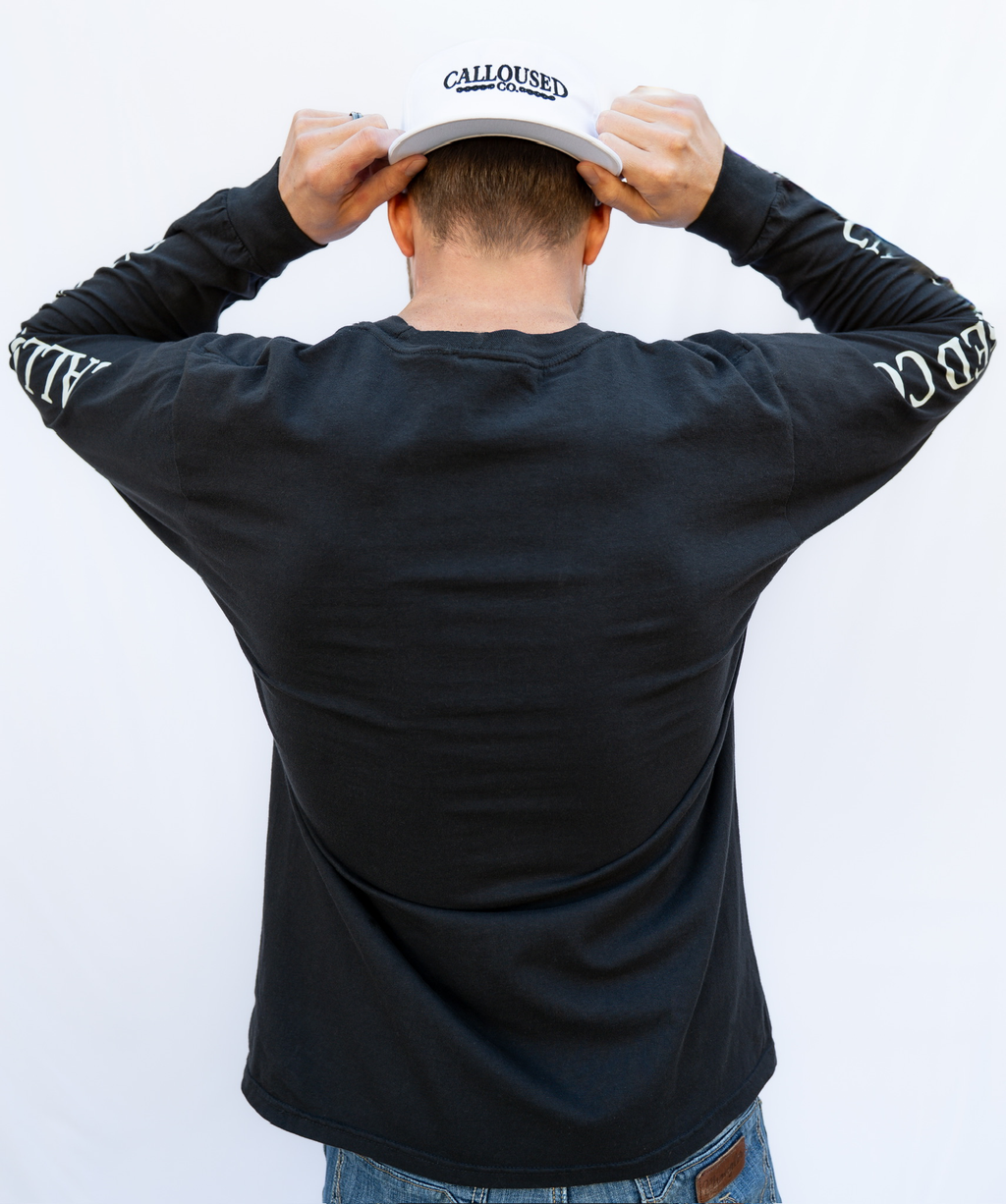 Person wearing a black long-sleeve shirt with 'Calloused Co' branding, holding a white cap with 'Calloused Co' logo.