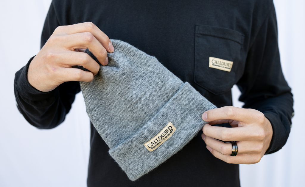 Person holding a gray beanie with 'Calloiused Co' branding against a white background
